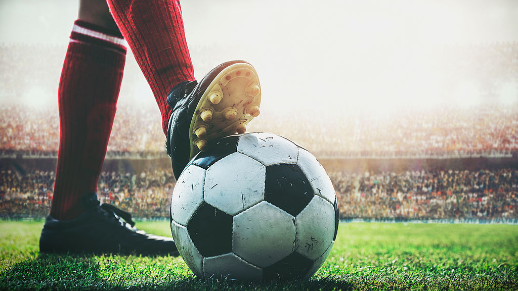 Der BdSt NRW appelliert an den Essener Stadtrat, kein weiteres Geld für den Ausbau des Stadions an der Hafenstraße bereitzustellen Stadion-Tribüne im Hintergrund zu sehen, Fußball auf Rasen groß im Mittelpunkt, Unterschenkel von Fußballspieler zu sehen mit Stollenschuh, einer auf dem Fußball