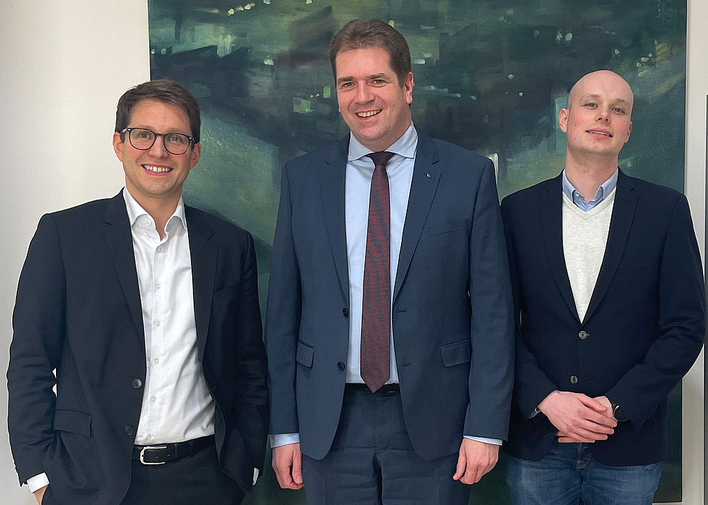 Der Finanzdezernent des Nordrhein-westfälischen und des Deutschen Städtetags Dr. Dominique Köppen (l.), der Vorsitzende des BdSt NRW, Rik Steinheuer, und BdSt-Haushaltsreferent Philipp Sprengel (r.). Der Finanzdezernent des Nordrhein-westfälischen und des Deutschen Städtetags Dr. Dominique Köppen (l.), der Vorsitzende des BdSt NRW, Rik Steinheuer, und BdSt-Haushaltsreferent Philipp Sprengel (r.) lächelnd vor einem angenehmen, abstrakten Bild, das mutmaßlich die Maße 2Meter mal Meter hat und in dunklem Blau sowie hellerem Grün gehalten ist. Die Farben sind spachtelartig aufgetragen.